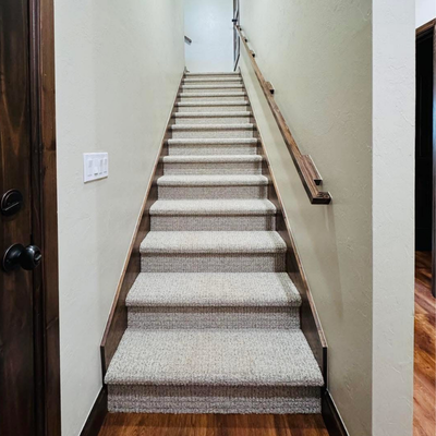 Carpeted staircase running upward between white walls, with wooden trim along both sides and a dark handrail mounted near the right wall, viewed from the bottom.